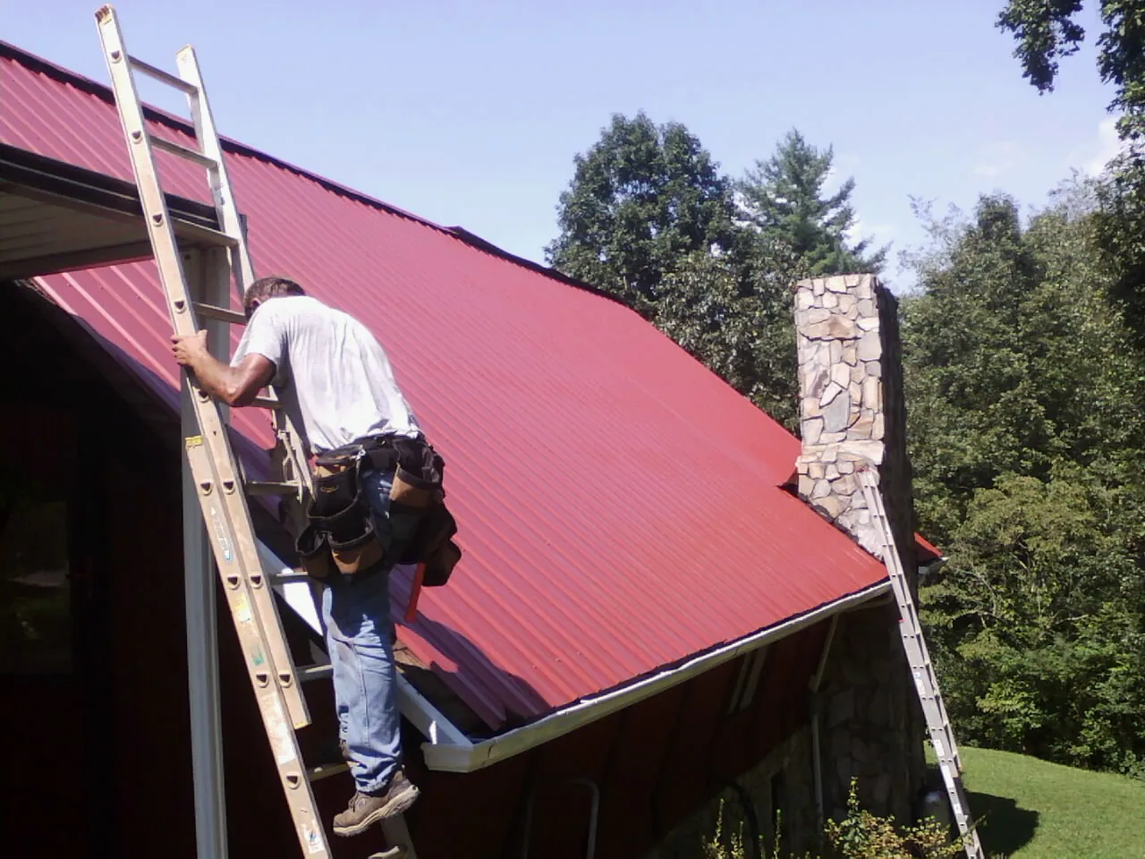 Roofer on a ladder beside a red metal roof and stone chimney — Soffit & Fascia Repair by Roofing Brothers in Amargosa Valley